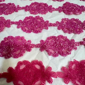 May include: Pink beaded trim featuring a floral design. The trim is made from small, round beads sewn together in a repeating pattern. This trim is ideal for adding a touch of elegance to any project.