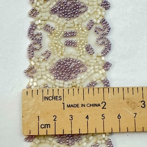 May include: Close-up of a beaded trim with a repeating floral design. The trim features lavender and off-white beads. A wooden ruler shows the trim is approximately 5 cm wide.
