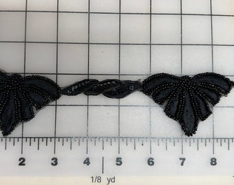 Pode incluir: Trimming preto com contas com um design de folha. O trimming mede aproximadamente 1/4 de jarda de comprimento e é mostrado em uma superfície quadriculada.