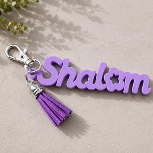 May include: A purple keyring featuring the word Shalom in a cursive font with a small flower cutout in the letter o. The keyring includes a silver metal lobster clasp and a matching purple suede tassel attached by a silver metal cap.