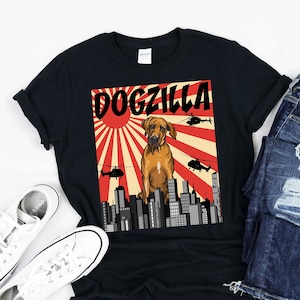 以下が含まれることがあります： 犬が「DOGZILLA」という文字の上に立っている、赤と白の昇る太陽を背景にした茶色の犬のグラフィックが描かれた黒いTシャツ。犬はヘリコプターが頭上を飛んでいる都市の風景の前に立っている