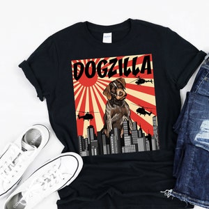 May include: Black t-shirt with a graphic of a brown and white dog standing on a city skyline with the text "DOGZILLA" above the dog. The graphic is in a retro style with a red sun and rays behind the dog.