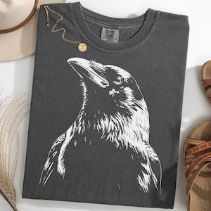 Puede incluir: Camiseta gris oscuro con una ilustración blanca detallada de la cabeza de un cuervo. La camiseta está doblada, con un collar dorado y un par de sandalias marrones. Un sombrero beige y hierba seca también están en la imagen.