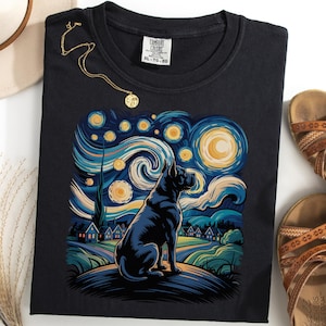 May include: Black t-shirt with a dog illustration in the style of Van Gogh's 'Starry Night'. The design uses blues, yellows, and greens. A gold necklace and brown sandals are also visible. The t-shirt is XL-TG size.