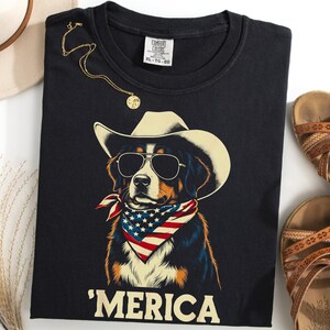 May include: A black t-shirt featuring a graphic of a Bernese Mountain Dog wearing a cowboy hat, sunglasses, and an American flag bandana. The text 'MERICA is printed in large cream-colored letters below the dog. The shirt has a Comfort Colors label.