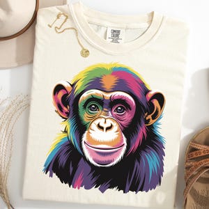Pode incluir: Camiseta creme com uma ilustração vibrante e colorida do rosto de um chimpanzé. A obra de arte usa um espectro de cores, incluindo amarelo, verde, roxo e rosa. A camisa é combinada com um colar de ouro, um chapéu e sandálias.