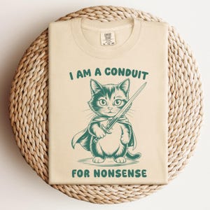 May include: A beige t-shirt with a green illustration of a cat holding a sword. The text "I am a conduit for nonsense" is printed on the shirt.
