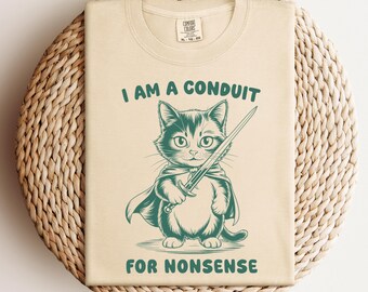 May include: A beige t-shirt featuring a green illustration of a cat holding a sword. The text "I am a conduit for nonsense" is printed on the shirt.