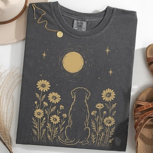 Op de afbeelding: Donkergrijs T-shirt met een gouden illustratie van een hond, maan en madeliefjes. Een gouden ketting en bruine sandalen zijn ook zichtbaar. Het T-shirt is gemaakt van zacht materiaal.