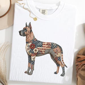 Può includere: T-shirt bianca con un disegno di cane alano. Il cane è decorato con un motivo patchwork della bandiera americana, fiori e altri elementi patriottici. La maglietta ha un'etichetta Comfort Colors.