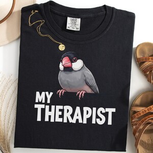 Op de afbeelding: Een zwart t-shirt met een afbeelding van een grijze rijstvogel met een roze snavel en de witte tekst "MY THERAPIST" onder de vogel gedrukt. Het shirt heeft een Comfort Colors merklabel bij de halslijn met de maat XL-TG-XG.