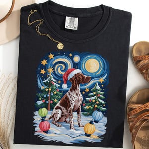 以下が含まれることがあります： サンタ帽をかぶったジャーマン・ショートヘアード・ポインターが描かれた黒いTシャツ。星空の夜景、クリスマスツリー、カラフルなライトが背景に。金のネックレスと茶色のサンダルも見える。