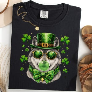 May include: Black t-shirt with a wolf design wearing a green top hat, sunglasses, and bow tie, with a green bubble and shamrocks. A gold necklace and brown sandals are also visible.