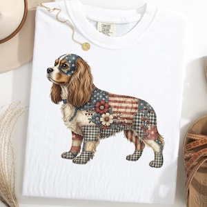 Op de afbeelding: Wit T-shirt met een Cavalier King Charles Spaniel ontwerp. De vacht van de hond is bedrukt met elementen van de Amerikaanse vlag, waaronder sterren en strepen, en bloemaccenten. Het shirt heeft een ronde hals en korte mouwen.
