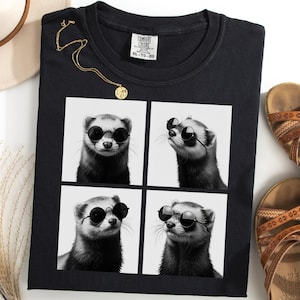 Puede incluir: Camiseta negra con un diseño de cuatro paneles de un hurón con gafas de sol. También se ven un collar dorado, un sombrero color crema y sandalias marrones. La camiseta está etiquetada como "Comfort Colors" y tiene una etiqueta de talla.
