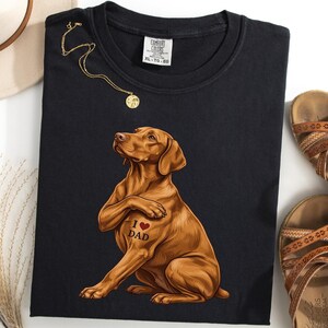 May include: Black t-shirt with a brown dog illustration and an "I ❤️ DAD" tattoo. A gold necklace, a hat, and brown sandals are also visible. The shirt is labelled "Comfort Colors" and size XL. The shirt is a casual wear item.