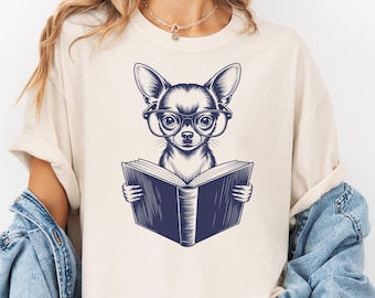 May include: Cream-colored t-shirt featuring a detailed illustration of a Chihuahua wearing glasses and reading a book. The dog is depicted in a navy blue ink style, with the book held open in its paws. The design is centered on the shirt.