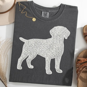 May include: Dark gray t-shirt featuring a white dog silhouette with a knitted texture design. The shirt is styled with a gold necklace, a beige hat, and brown sandals. A casual, everyday wear tee.