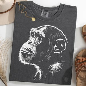 Pode incluir: T-shirt cinza escuro com uma ilustração de chimpanzé branco. A imagem inclui sandálias castanhas, um chapéu bege e um colar dourado. A t-shirt é feita de um material macio e confortável.