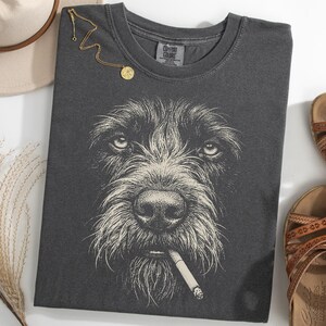 以下が含まれることがあります： タバコをくわえた犬のイラストが描かれた濃いグレーのTシャツ。ベージュの帽子、金のネックレス、茶色のサンダルがTシャツの周りに配置されています。