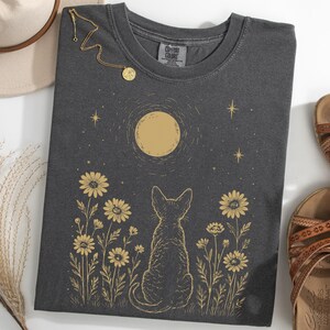 May include: Dark gray t-shirt with a cat silhouette gazing at a golden moon and stars, surrounded by golden daisies. A gold necklace and brown sandals are also visible. The shirt is a Comfort Colors brand.