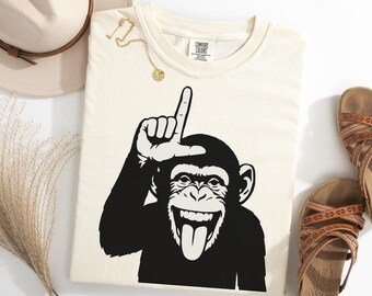 May include: Cream-colored t-shirt featuring a black and white graphic of a chimpanzee with its tongue out and giving the 'loser' gesture. The shirt is from Comfort Colors. Accessories include a gold necklace, a beige hat, and brown sandals.