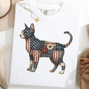 Könnte beinhalten: Weißes T-Shirt mit einem patriotischen Katzendesign. Die Katze ist ein Patchwork aus Elementen der amerikanischen Flagge, einschließlich Sterne, Streifen und floralen Akzenten. Das Shirt hat ein Comfort Colors-Etikett.