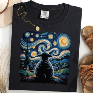 Puede incluir: Camiseta negra con una colorida ilustración de una oveja mirando un cielo estrellado, al estilo de Van Gogh. También se ven un collar dorado y sandalias marrones.