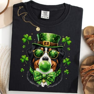 May include: Black t-shirt featuring a Bernese Mountain Dog wearing a green top hat, sunglasses, and bow tie, with a green bubble gum bubble. The design includes shamrocks. A gold necklace and sandals are also visible.