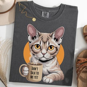 以下が含まれることがあります： コーヒーカップを持った猫のイラストが描かれた濃いグレーのTシャツ。猫は茶色と白の毛並みで、黄色い目をしています。オレンジ色の円が背景にあります。マグカップには「DON'T TALK TO ME YET」と書かれています。