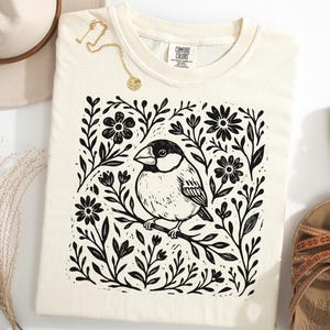 May include: Cream-coloured t-shirt with a black and white linocut print of a bird on a branch, framed by flowers and foliage. The shirt is styled with a hat, sandals, and a gold necklace.