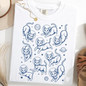 May include: White t-shirt with a navy blue design of cats wearing space helmets, alongside planets and stars. The cats are depicted in different poses, creating a fun and imaginative pattern. The shirt is a Comfort Colors brand.