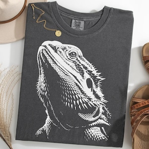 May include: Charcoal grey t-shirt with a white graphic of a bearded dragon's head. A gold necklace, a beige hat, and brown sandals are also visible. The t-shirt is made of a soft material. The shirt is a Comfort Colors brand.