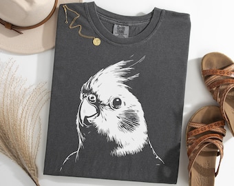May include: Dark gray Comfort Colors t-shirt featuring a large white illustration of a cockatiel's head. A gold necklace with a circular pendant rests on the shirt. Tan sandals and a beige hat are also in the image.