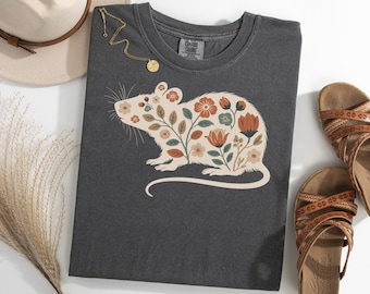 May include: Dark gray t-shirt featuring a floral rat design in cream, orange, and green. The shirt is styled with a hat, sandals, and a gold necklace. The design includes various flowers and leaves.