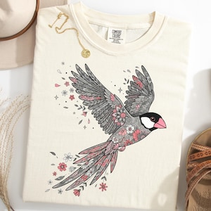 Puede incluir: Camiseta color crema con una ilustración detallada de un pájaro en vuelo. El pájaro tiene alas y plumas de la cola grises con detalles en rosa y flores. Se ven un collar dorado y un sombrero.