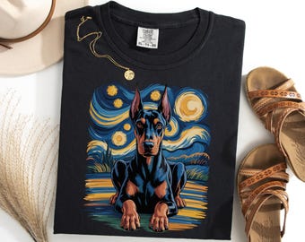 Puede incluir: Camiseta negra con una ilustración de Doberman Pinscher al estilo de "La noche estrellada" de Van Gogh. El perro está representado con un cielo nocturno azul y amarillo. La camiseta tiene una etiqueta Comfort Colors.