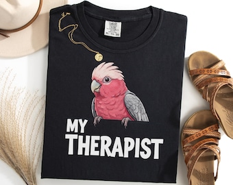 May include: A black t-shirt featuring a graphic of a pink and grey Galah cockatoo perched on the edge of the text "MY THERAPIST" printed in white. The shirt has a Comfort Colors brand label at the neckline and is size XL.