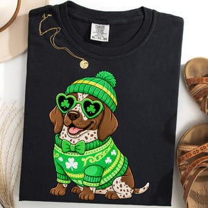 May include: Black t-shirt featuring a cartoon dog wearing a green and yellow hat, heart-shaped sunglasses, and a green sweater with shamrocks. The shirt also has a gold necklace and is next to a pair of brown sandals and a cream-colored hat.