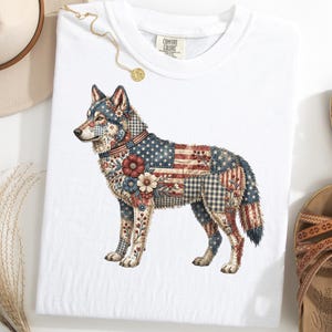 May include: White t-shirt featuring a detailed illustration of a wolf. The wolf's body is adorned with a patchwork design incorporating the American flag, floral patterns, and various textures. The shirt has a crew neck and short sleeves.