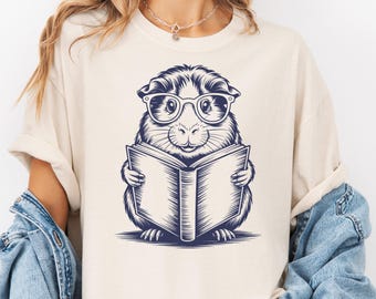 May include: Cream-colored t-shirt featuring a navy blue illustration of a guinea pig wearing glasses and reading a book. The guinea pig is the central focus, with detailed shading and a friendly expression. The design is simple yet charming.
