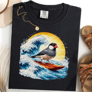 Può includere: Maglietta nera con un uccello che fa surf su un'onda e un sole giallo. Sono visibili anche una collana d'oro, un cappello di paglia e sandali marroni.