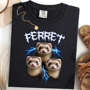 Puede incluir: Camiseta negra con la palabra "FERRET" en blanco, con letras góticas, y tres retratos de hurones con rayos azules. También se ven un collar dorado, un sombrero beige y sandalias marrones.