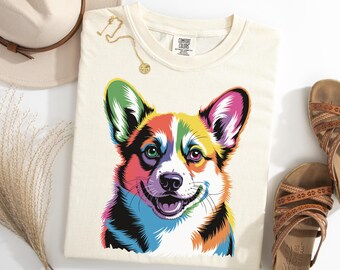 Pode incluir: Camiseta creme com uma ilustração vibrante em estilo pop art de um cão Corgi sorrindo. A pelagem do cão é representada em um arco-íris de cores. A camiseta é da Comfort Colors.