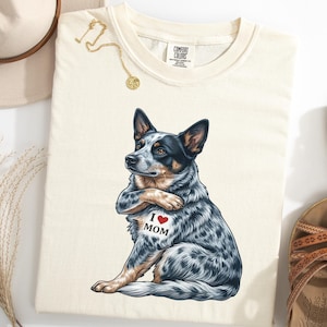 May include: Cream-coloured Comfort Colors t-shirt with a blue heeler dog illustration and the text "I ❤️ MOM". The dog is sitting and looking to the side. A gold necklace and a hat are visible.
