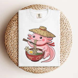 Puede incluir: Camiseta blanca con un axolotl rosa de dibujos animados que lleva un sombrero de paja y come fideos ramen de un cuenco. El texto "COMFORT COLORS" está en la camiseta.
