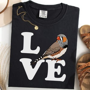 Könnte beinhalten: Schwarzes T-Shirt mit dem Wort "LOVE" in Weiß und einer farbenfrohen Illustration eines Zebrafinken. Eine goldene Halskette und braune Sandalen sind ebenfalls zu sehen.