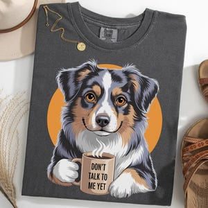 May include: Dark gray t-shirt featuring a colorful illustration of an Australian Shepherd dog holding a coffee mug. The mug reads "DON'T TALK TO ME YET". The dog is set against an orange circle. A gold necklace, hat, and sandals are also in the image.
