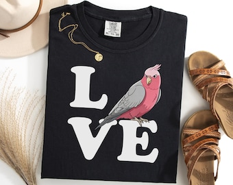 May include: Black t-shirt with the word "LOVE" in white block letters. A pink and gray bird graphic is perched on the "VE". A gold necklace and brown sandals are also in the image. The shirt is a Comfort Colors XL-TG.
