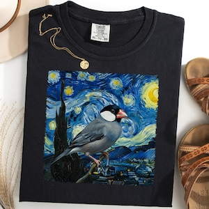 May include: Black t-shirt featuring a bird design set against a starry night background, reminiscent of Van Gogh's style. The shirt is accompanied by a gold necklace, sandals, and a hat.